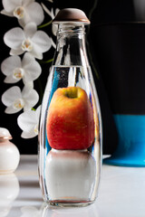 still life distorted apple behind glass bottle with water, on marble and white orchids in the background