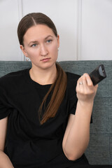 Bored young woman sitting on a grey sofa in her living room, holding a remote control and changing...