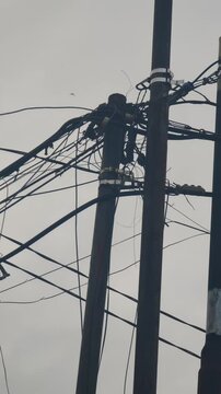Portrait-format video showing a chaotic tangle of electrical and telephone cables hanging across utility poles in an urban neighborhood. The disorganized wiring system represents infrastructure