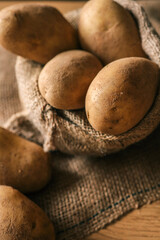 Fresh raw potatoes in a rustic burlap sack on a wooden surface. Organic vegetables displayed in a...