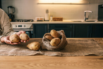 Hands holding and arranging fresh potatoes in a burlap sack on a wooden table in a cozy kitchen....
