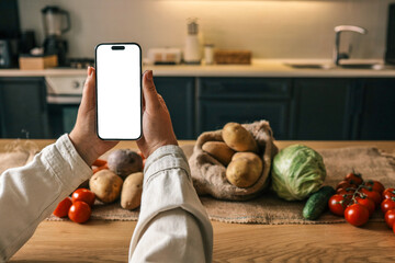 Person holding a smartphone with blank white screen over a rustic kitchen table filled with fresh vegetables: potatoes, tomatoes, cabbage, cucumber. Mockup concept with natural lighting