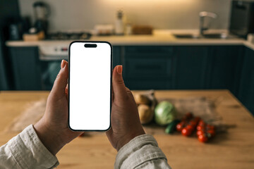 Person holding a smartphone with blank white screen over a rustic kitchen table filled with fresh vegetables: potatoes, tomatoes, cabbage, cucumber. Mockup concept with natural lighting