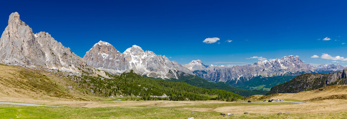 A sweeping panoramic view from Passo di Giau, showcasing rugged Dolomite peaks, verdant valleys, and a clear blue sky, with a winding road below.