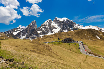The winding road of Passo di Giau leads to a mountain lodge, with the snow-capped peak of Antelao dominating the background under a vibrant blue sky.