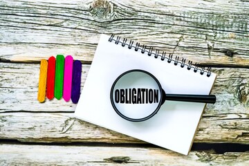 The word OBLIGATION on a notebook is seen using a magnifying glass isolated on a wooden table.