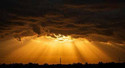 Dramatic god rays and golden sunbeams bursting through dark storm clouds at sunset. A spiritual, religious background symbolizing hope, heaven, and faith over a landscape silhouette. Epic sky.