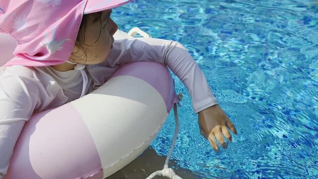 Close-up shot of a happy 5-year-old mixed Korean girl wearing a pink hat and floating on an inflatable ring, enjoying summer play in a bright blue swimming pool. - Powered by Adobe