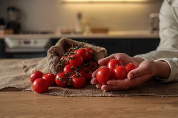 Hands holding fresh red tomatoes next to a burlap sack filled with vine tomatoes on a rustic wooden table in a cozy kitchen