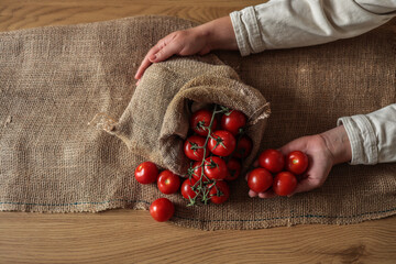 Hands holding fresh red tomatoes next to a burlap sack filled with vine tomatoes on a rustic wooden...