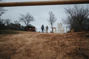 A pair of friends exploring a serene natural park, surrounded by bare trees and overcast skies. The...