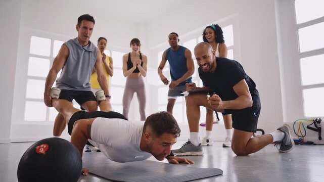 Fitness instructor supporting athlete's legs while he performs push-ups, demonstrating teamwork and motivation in gym environment with supportive classmates cheering