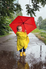 Joyful Child in a Yellow Raincoat Holding a Bright Red Umbrella While Splashing in Rainwater on a...
