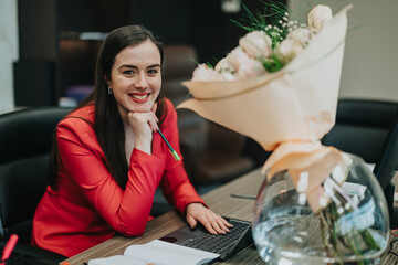 Professional woman in red suit in an office environment, seated at a desk with flowers and a...
