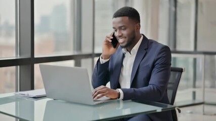 Confident businessman talking on smartphone while working on laptop at desk - Powered by Adobe