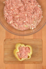 A single bell pepper on a wooden board is being stuffed with a mixture of ground meat, rice, and onions, with a bowl of the remaining mixture visible above it