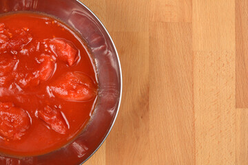 A close-up, overhead view shows a metal bowl filled with whole peeled tomatoes in rich red sauce, positioned on the left side of a wooden surface, with ample copy space on the right