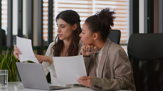 Multiethnic diverse girls ladies women business partners coworkers African American Caucasian female teamwork review documents using laptop analyze information corporate office smiling businesswomen