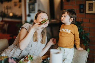 A mother and young child share a lighthearted moment eating food with melted cheese. They interact...