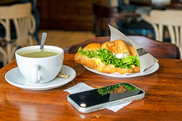 hot croissant with tuna, egg and salad near the phone with ginger tea. Lunch snack
