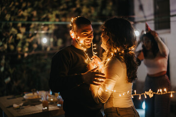 A joyful couple smiles and embraces, wrapped in string lights, celebrating outdoors at night.
