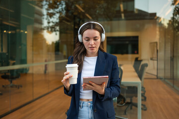 Businesswoman wearing headphones using digital tablet and holding coffee outside office building