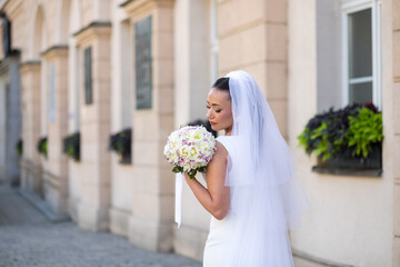 Elegant bride in white wedding dress holding a bouquet, standing by a historic building with floral...