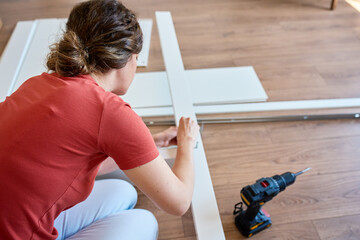 Woman sits on wooden floor and assembling furniture in new home, fastens parts of white bed frame....