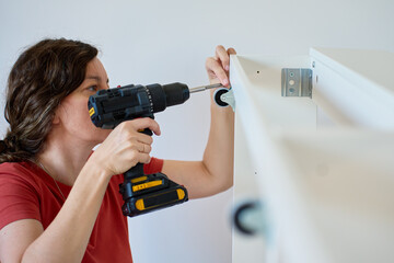 Woman using cordless drill to screw part into white bed frame. Female assembling furniture in new...