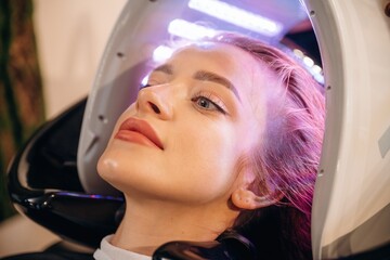 Pink or velvet color. Woman is in a beauty salon with LED hair growth helmet