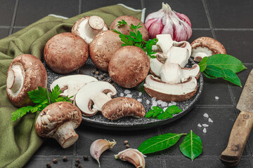 Fresh brown mushrooms with garlic, herbs and rustic knife on black tile kitchen surface