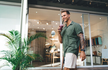 Young man sipping smoothie in front of boutique windows, portraying connected leisure, mobile...
