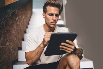 Focused man holding stylus and interacting with tablet while seated on staircase, showing detailed...