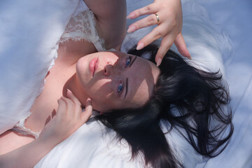 curvy woman with blue eyes lies in soft daylight on white bedding, framed by black hair and lace...
