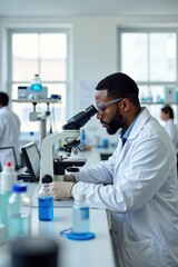 A black man using a microscope in a bright laboratory filled with chemical bottles and scientific instruments. The background has white walls and large windows
