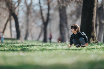 A young child wearing a dark sweater plays in the grass of a park surrounded by trees on a sunny...