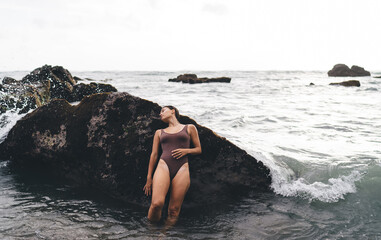 Young woman stands against sea-splashed rock, head tilted, eyes closed, embodying total digital disconnection and surrender to nature’s rhythm, raw emotion, and timeless presence.