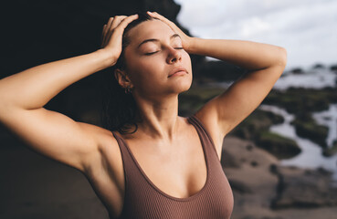 Close-up portrait of young woman with eyes closed and hands in hair, radiating peaceful introspection, physical awareness and deep mental calm far from social feed overload.