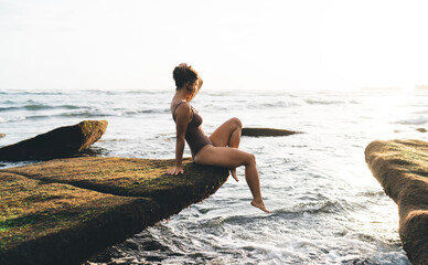 Young woman sits on mossy ledge above waves, arm raised, face shaded, revealing moment of...