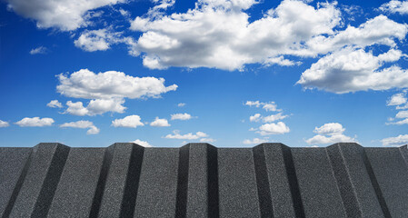 A low-angle view of a dark, textured corrugated metal fence against a vibrant blue sky with fluffy...