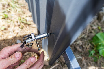 A person's hands using a riveter to fasten a black corrugated metal sheet, for a fence or structure.