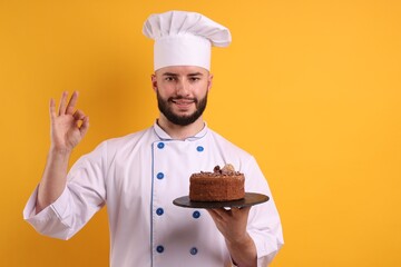 Happy confectioner in uniform holding chocolate cake and showing ok gesture on orange background