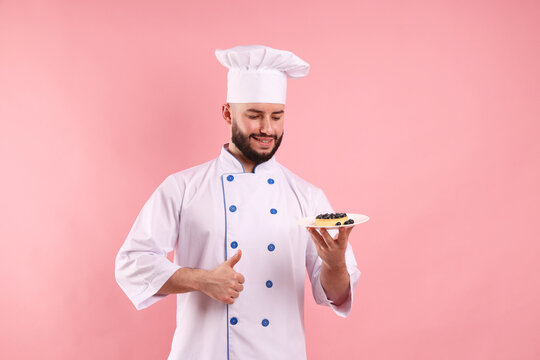 Happy confectioner in uniform holding tart with blueberries and showing thumbs up on pink background