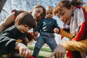 Two mothers bond with their young children during an engaging play moment in a sunny park, sharing...