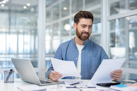 A smiling businessman reviews documents at his desk in a bright, modern office setting. - Powered by Adobe