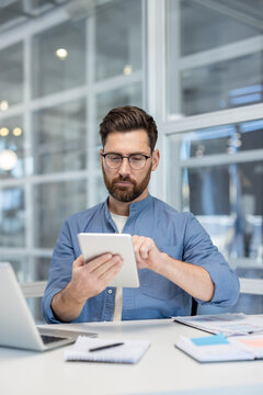 Fototapeta A man in glasses uses a tablet at his desk in a modern office environment.