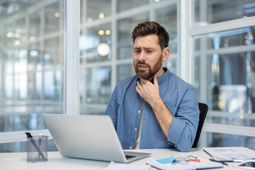 A man in a blue shirt looks concerned while working on his laptop, touching his throat.