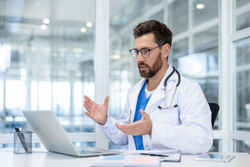 A doctor in a white coat sits at a desk, gesturing while on a video call using a laptop, discussing...