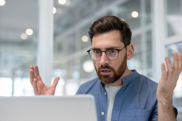 A man with glasses is surprised by something he sees on his laptop. He has his hands up in...