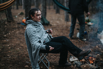 A young woman enjoys the outdoors sitting by a campfire, wrapped in a blanket, with hammocks and...